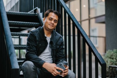 Genç bir Hindu adam şehir caddesinde metal bir merdivende oturuyor ve telefonunu kontrol ederken sabah kahvesinin tadını çıkarıyor. Turizm ve şehir yaşam tarzı kavramı.