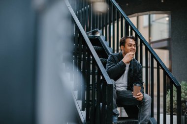 Neşeli genç bir Hindu adam elinde telefonla şehrin bir caddesinde metal bir merdivende oturuyor. Turizm ve şehir yaşam tarzı kavramı.