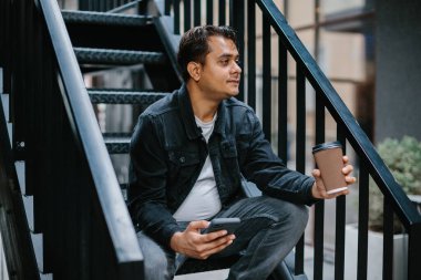 Genç bir Hindu adam şehir caddesinde metal bir merdivende oturuyor ve telefonunu kontrol ederken sabah kahvesinin tadını çıkarıyor. Turizm ve şehir yaşam tarzı kavramı.