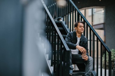 Neşeli genç bir Hindu adam elinde telefonla şehrin bir caddesinde metal bir merdivende oturuyor. Turizm ve şehir yaşam tarzı kavramı.