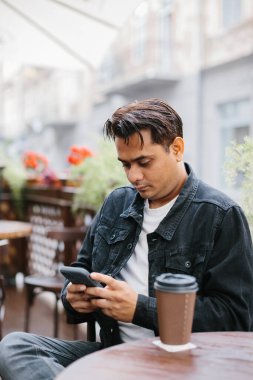 Neşeli genç Hintli bir adam bir kafenin terasında kahve içiyor ve sosyal ağlara takılıyor ya da telefonda sohbet ediyor..