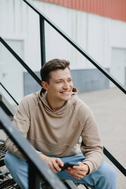 Genç bir adam sokağın metal merdivenlerinde oturuyor, telefonu elinde tutuyor..