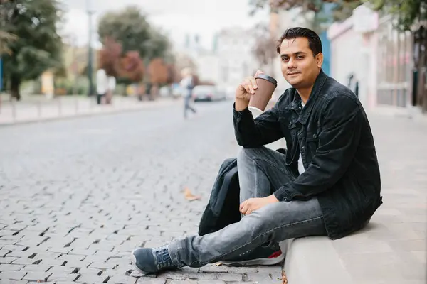 Gülümseyen bir Hindu adam bir şehir caddesinde yerde oturuyor ve sabah kahvesinin tadını çıkarıyor. Turizm ve şehir yaşam tarzı kavramı.
