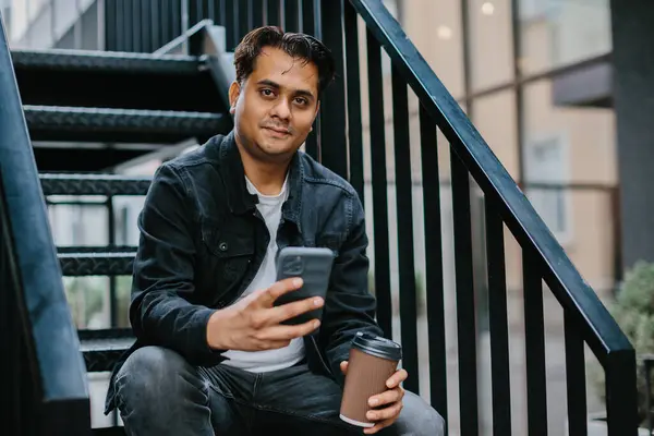 Genç bir Hindu adam şehir caddesinde metal bir merdivende oturuyor ve telefonunu kontrol ederken sabah kahvesinin tadını çıkarıyor. Turizm ve şehir yaşam tarzı kavramı.