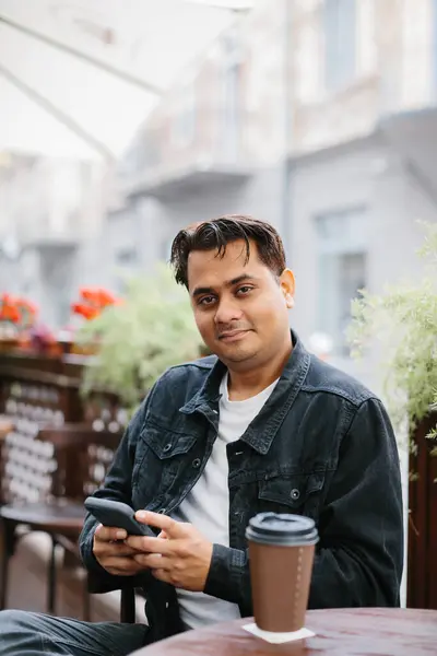A cheerful young Indian man is drinking coffee on the terrace of a cafe and browsing social networks or chatting on his phone.