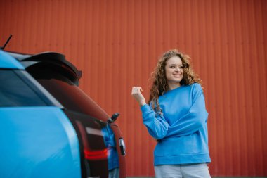 Havalı genç bir kadın park yerinde elektrikli arabasının yanında poz veriyor..