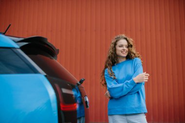 Havalı genç bir kadın park yerinde elektrikli arabasının yanında poz veriyor..