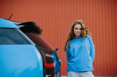 Havalı genç bir kadın park yerinde elektrikli arabasının yanında duruyor..