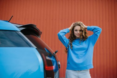 Havalı genç bir kadın park yerinde elektrikli arabasının yanında poz veriyor..