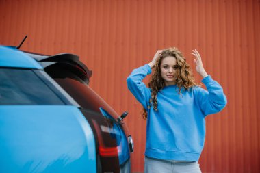 Havalı genç bir kadın park yerinde elektrikli arabasının yanında poz veriyor..