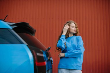 Havalı genç bir kadın park yerinde elektrikli arabasının yanında poz veriyor..