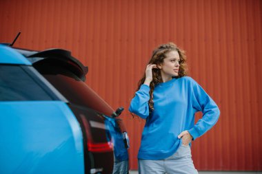 Havalı genç bir kadın park yerinde elektrikli arabasının yanında poz veriyor..