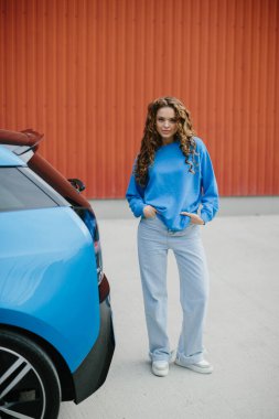 Havalı genç bir kadın park yerinde elektrikli arabasının yanında duruyor..