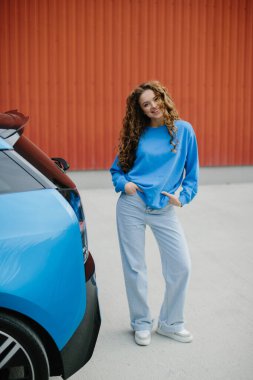 Havalı genç bir kadın park yerinde elektrikli arabasının yanında duruyor..