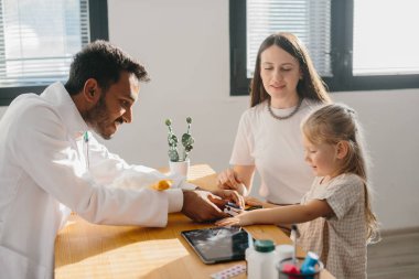 Genç bir çocuk doktoru küçük bir hastanın nabzını ölçer. Anaokulu kızı doktor randevusunda olan bir anne..