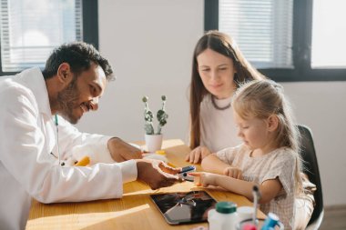 Genç bir çocuk doktoru küçük bir hastanın nabzını ölçer. Anaokulu kızı doktor randevusunda olan bir anne..