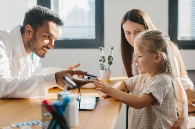 Genç bir çocuk doktoru küçük bir hastanın nabzını ölçer. Anaokulu kızı doktor randevusunda olan bir anne..