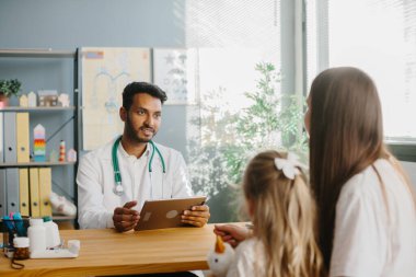 Elinde tabletle genç bir erkek çocuk doktoru küçük kızına bir anneye danışıyor. Çocuk doktoru randevusu konsepti.