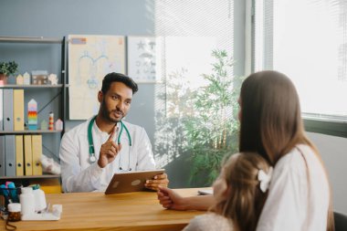 Elinde tabletle genç bir erkek çocuk doktoru küçük kızına bir anneye danışıyor. Çocuk doktoru randevusu konsepti.