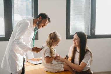 Erkek çocuk doktoru steteskopla küçük tatlı bir kızı muayene ediyor.