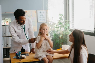 Anne ve çocuk aile hekimi ile görüşüyor. Steteskopla gülümseyen çocuk doktoru küçük kızın ciğerlerini kontrol ediyor..