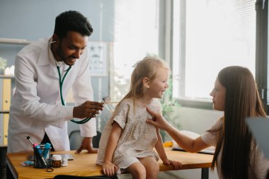 Anne ve çocuk aile hekimi ile görüşüyor. Steteskopla gülümseyen çocuk doktoru küçük kızın ciğerlerini kontrol ediyor..
