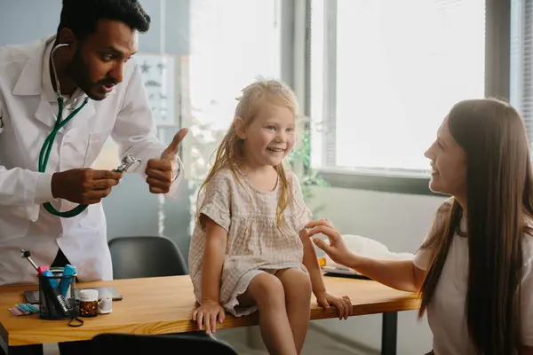 Anne ve çocuk aile hekimi ile görüşüyor. Steteskopla gülümseyen çocuk doktoru küçük kızın ciğerlerini kontrol ediyor..