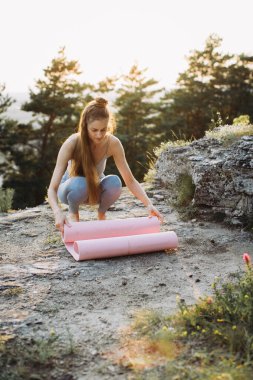 Güzel bir dağ yerinde yapılan çalışmadan sonra spor kıyafeti giymiş genç bir kadın yoga minderini kaldırıyor..