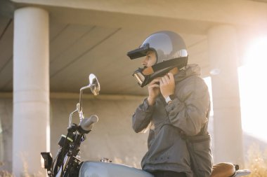 Motorcunun biri motosikletin üstünde otururken kask takar.