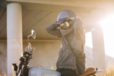 Motorcunun biri motosikletin üstünde otururken kask takar.