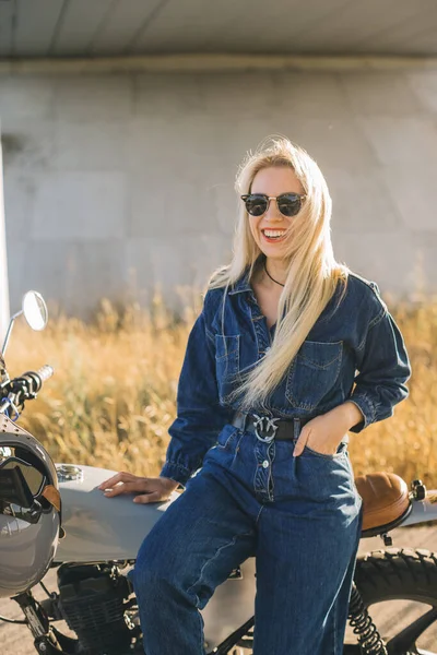 A young blonde woman in denim clothes and sunglasses poses on a motorcycle at sunset.