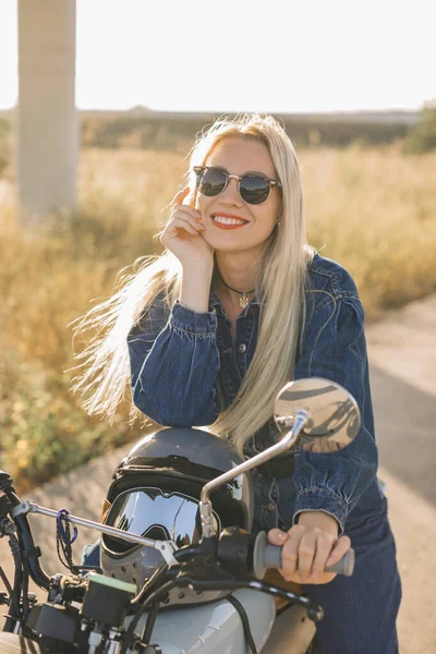 A young blonde woman in denim clothes and sunglasses poses on a motorcycle at sunset.
