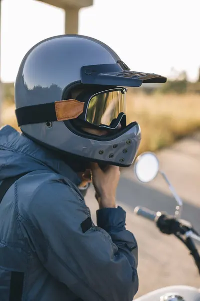A motorcycle rider fastens his helmet while sitting on a motorcycle on the road against the sunset.