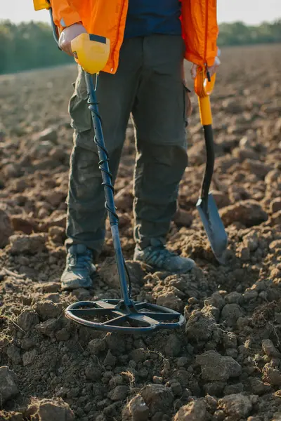 A coil of a metal detector shovel and the legs of a person looking for treasure against the background of a plowed field.