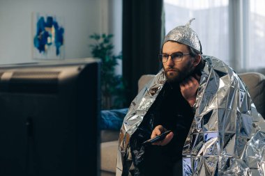 Folyo şapka takan ve folyoya sarılı genç bir adam elinde uzaktan kumandayla televizyonun önündeki koltukta oturuyor..
