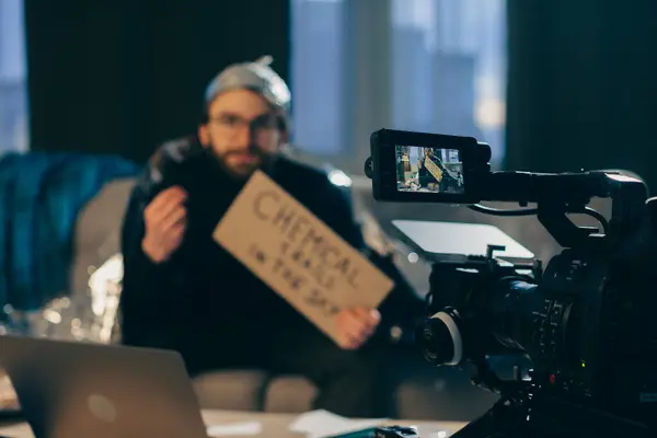 陰謀論者は カメラに疑似科学的なビデオを撮影する スズキホイルの帽子と手のサインの男は カメラの前にソファに座っています — ストック写真
