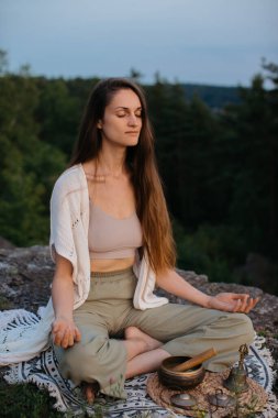 A young beautiful brunette woman meditates in the mountains on the background of a pine forest in the rays of the setting sun. clipart