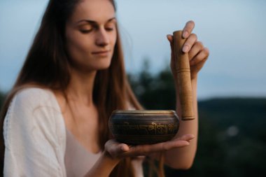 Genç esmer kadın gün batımında dağlarda şarkı söyleyen bir kâseyle meditasyon yapıyor..