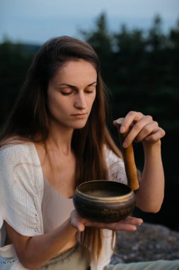 Genç esmer kadın gün batımında dağlarda şarkı söyleyen bir kâseyle meditasyon yapıyor..