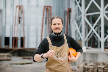 Koruyucu miğferli, gözlüklü ve tulumlu bir müteahhit tahıl ambarının arka planında duruyor..