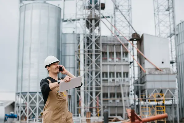 An engineer works in an industrial area on the background of a silo with grain.