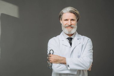 Elinde fallendoskop olan beyaz önlüklü kıdemli bir doktor gri arka planda poz veriyor. Sağlık ve Tıp Konsepti.