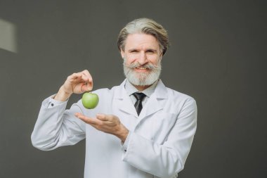 Beyaz önlüklü, elinde yeşil elmayla gri arka planda poz veren yakışıklı bir kıdemli doktor. Sağlık ve Tıp Konsepti.