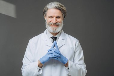 Gri arka planda beyaz önlüklü, neşeli, kıdemli bir doktorun portresi..