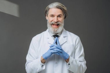 Gri arka planda beyaz önlüklü, neşeli, kıdemli bir doktorun portresi..