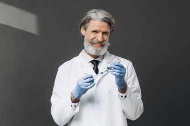 Beyaz önlüklü yaşlı doktor elinde şırıngayla gri arka planda izole edilmiş..