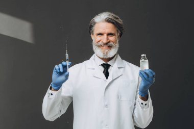 Beyaz önlüklü yaşlı doktor elinde şırınga ve şişeyle gri arka planda izole edilmiş..