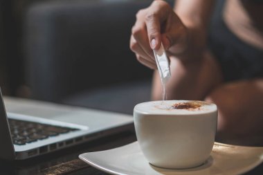 Kafe 'deki ahşap masada laptoplu bir fincan sıcak latte kahve tutan bir elin yakın plan görüntüsü.