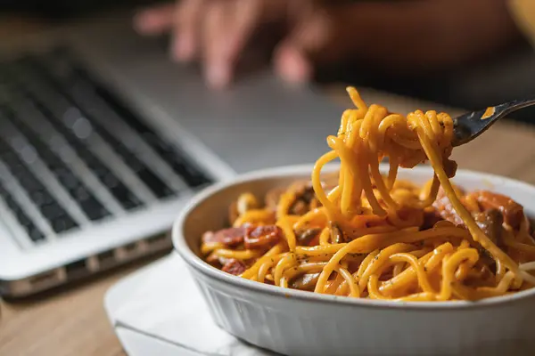 Eating spaghetti while working on laptop at home. Unhealthy food during conference call, meeting.