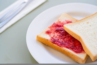 Close-up of two slices of white bread on a white plate, one with strawberry jam spread on it. A fork and knife are placed on a napkin in the background. clipart